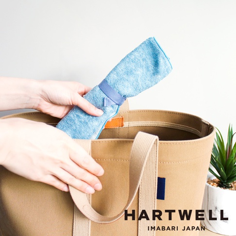 雙手將藍色毛巾捲起，用藍色綁帶固定，放入卡其色帆布托特包中。