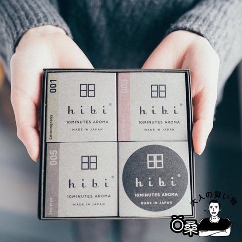 雙手捧著一盒由四個小紙盒組成的香氛火柴禮盒。禮盒外觀為灰褐色，每個小紙盒上印有「hibi」品牌標誌、數字編號（001, 003, 005）及香氛名稱（Lemongrass, Geranium, Tea tree），其中一個圓形紙盒印有「hibi」標誌。
