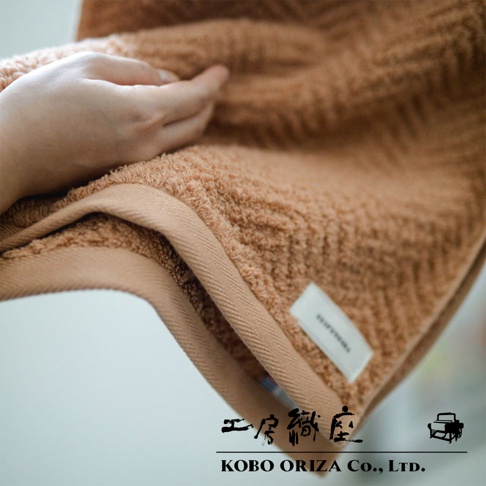 一隻手輕柔地觸碰著一件淺棕色、具有人字紋理的有機棉浴巾。浴巾邊緣有著米色的滾邊，上面縫有白色的小標籤。