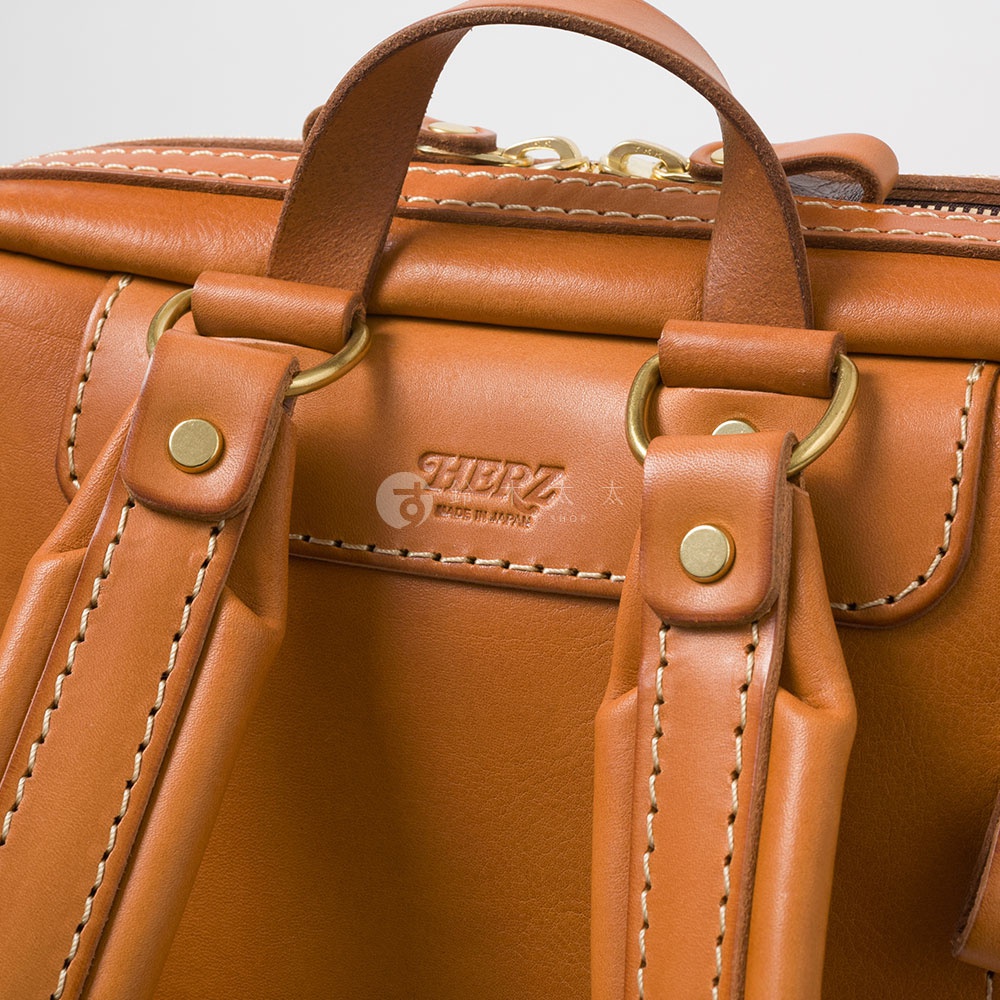 職人手作皮革軟皮商務後背包 R-28，近拍特寫，展現溫潤的棕色皮革材質、精緻的縫線細節、黃銅色五金配件，以及壓印的品牌標誌。