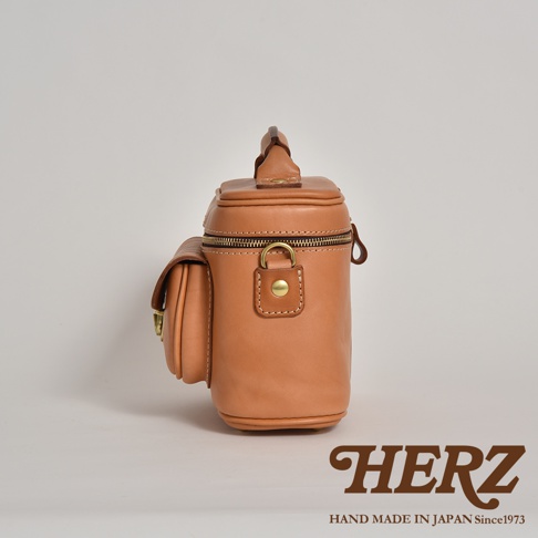 職人手作皮革軟皮相機包 N-6，呈現溫潤的焦糖色調。包身為圓筒狀，頂部有提把，側邊設有一個翻蓋式小口袋，以銅釦固定。主袋口為拉鍊設計，搭配復古風格的拉鍊頭和金屬D型環。整體採用厚實的皮革材質，展現精緻的手工縫線。