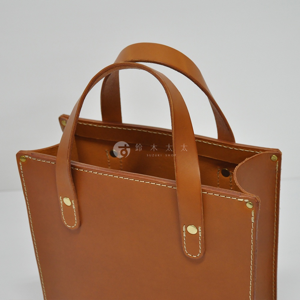 近拍特寫展示一個 tan 色皮革製成的直式托特包，包包有兩條皮革提把，以白色縫線和金色鉚釘裝飾，整體線條簡潔。