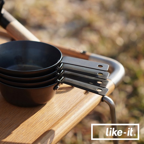 一疊黑色金屬材質的萬能調理把手鐵杯，帶有長柄，置於木製檯面上，背景模糊，顯示戶外環境。