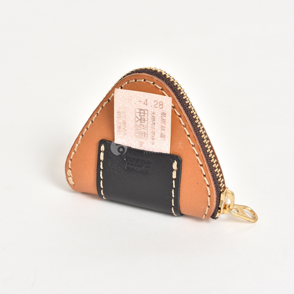 日本HERZ職人手作皮革三角飯糰造型零錢包，以淺棕色皮革為主體，搭配黑色皮革外層口袋，口袋上印有HERZ品牌壓印，黑色外袋中插入一張寫有弘南鐵道中央弘前站日期的車票，整體採粗縫線設計，側面配有金色拉鍊，呈現簡約且獨特的精緻工藝風格。