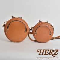 兩個職人手作皮革小圓包，皆為天然棕色皮革，圓形設計，頂部有皮革蝴蝶結裝飾，正面有拉鍊開合，搭配金屬拉鍊和皮革拉頭。