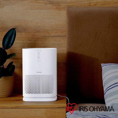 白色直立式空氣清淨機，正面有黑色網格狀出風口，上方有控制面板及品牌標誌，擺放在木質床頭櫃上，旁有綠葉植物及床鋪。