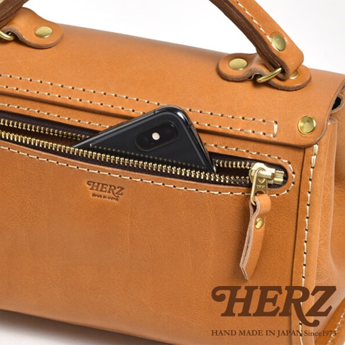 職人手作皮革迷你手提包，顏色為棕褐色。包包採用厚實皮革製成，縫線精緻，帶有拉鍊前袋，可放入手機。包包配有提把和金屬扣環，質感扎實。