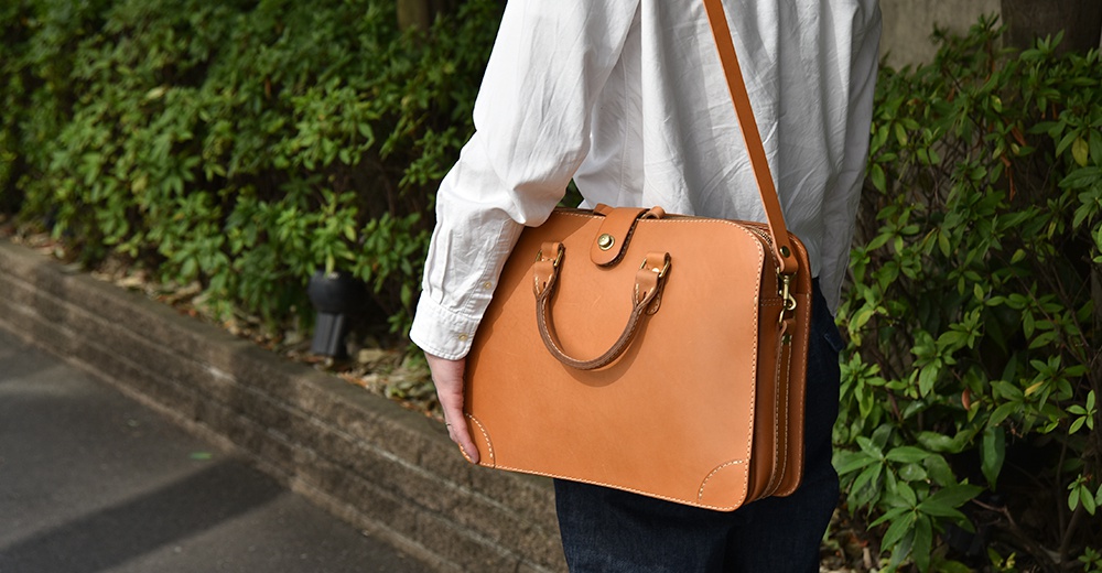 淺棕色皮革製成的手提公事包，有兩個頂部提把、可拆卸肩帶和磁扣開合。