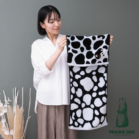 一位留著黑色短髮的女性穿著白色襯衫與棕色寬褲，手持一條黑白相間的斑點圖案毛巾，毛巾上方邊緣繡有一隻精緻的貓咪臉部特徵，下方則有 PRAIRIE DOG 的品牌標誌與官網網址。