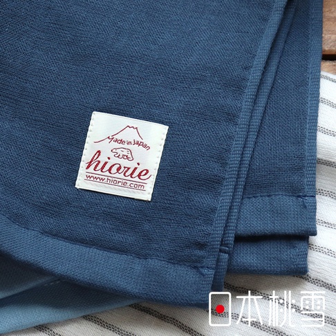 藍色紗布毛巾，表面紋理清晰，左側有標籤，標示「made in Japan」和「hiorie」，材質輕薄透氣，邊緣有縫製。
