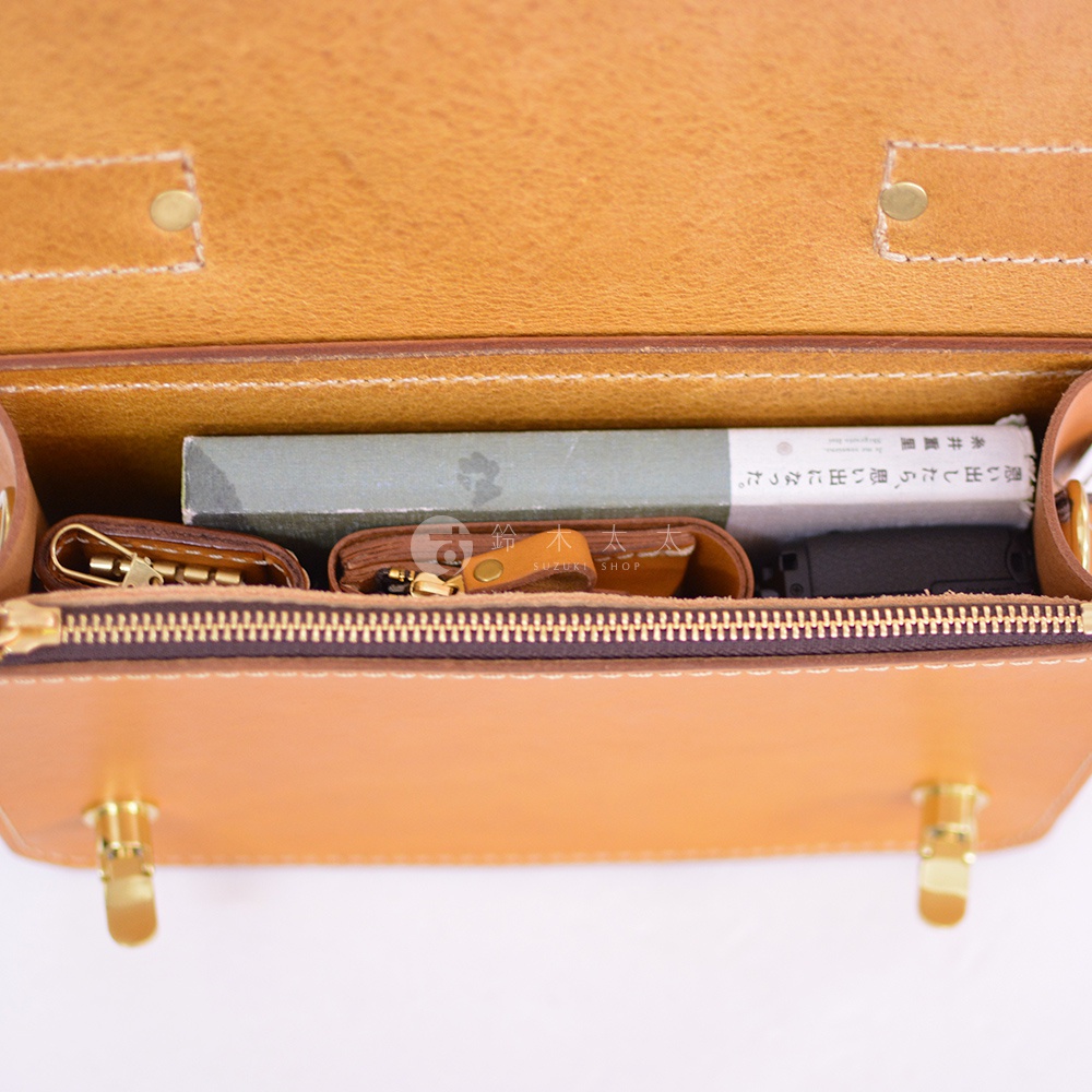 職人手作皮革經典兩用雙扣迷你單肩包，以自然色澤的皮革製成，包內展示有鑰匙包、皮夾、書本和黑色物品。