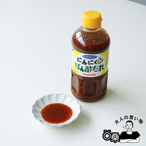 一瓶帶有黃色瓶蓋的透明塑膠瓶裝朝紫大蒜酸橘醋風味調味醬，瓶身標籤寫有日文品名，左側擺放一個盛裝著少量深褐色醬汁的白色波浪花邊小碟，整體置於純白色的背景桌面上，右下方帶有一個卡通男性人物的插畫標誌。