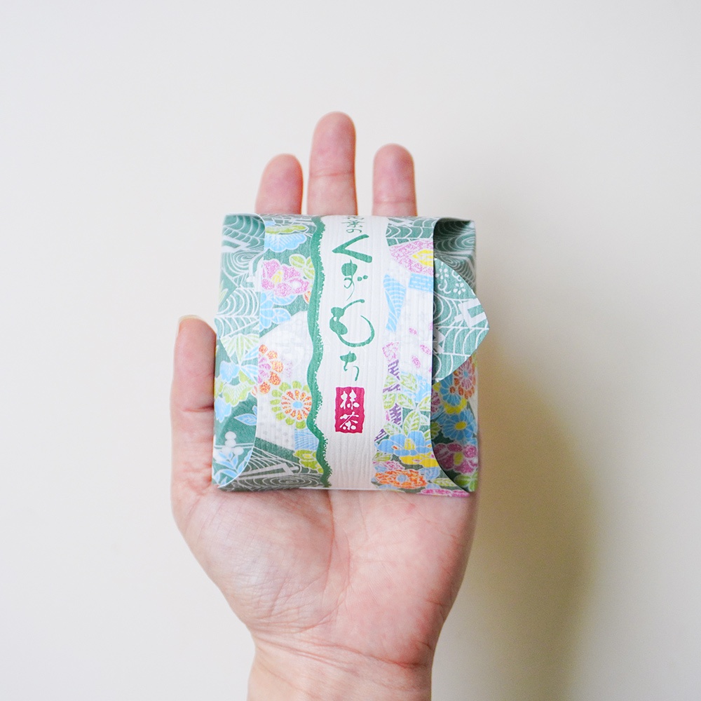 一隻手掌平托著一個正方形的小點心，點心外層以印有精緻花卉圖案的日式友禪染風格包裝紙包裹，中間貼有一條印有綠色草寫書法字樣抹茶葛餅的標籤紙。