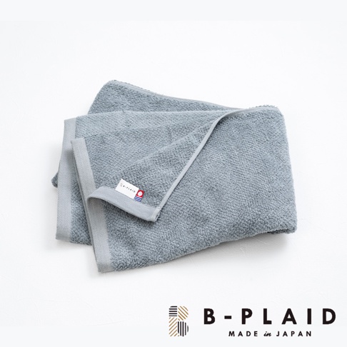 一條灰藍色、觸感柔軟的薄手鱗紋浴巾，帶有灰色的邊緣和一個標有「B-PLAID」及日本國旗圖案的白色標籤。