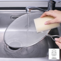 一只手正在用黃色海綿清潔一個銀色的金屬網狀透視防噴濾網，濾網置於水龍頭下方、流理臺水槽中，濾網邊緣有金屬框和手柄。