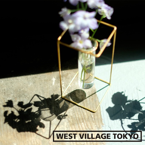 黃銅四角線條構成的簡約花器，內置玻璃試管，盛有少量清水與綠色植物莖葉，上方擺放著淡紫色小花。