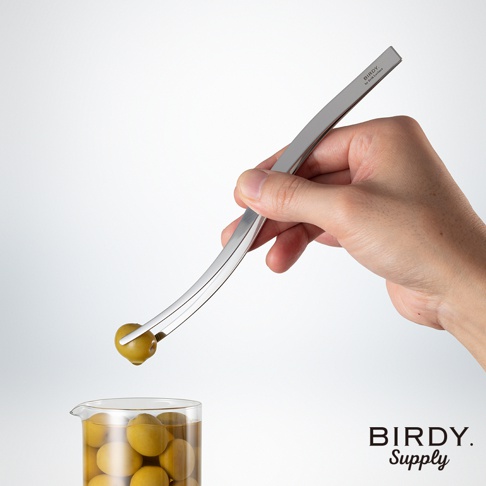 銀色金屬材質的不沾桌調酒專用裝飾夾，其彎曲的夾臂夾著一顆綠色橄欖，正要放入裝有橄欖的玻璃杯中。