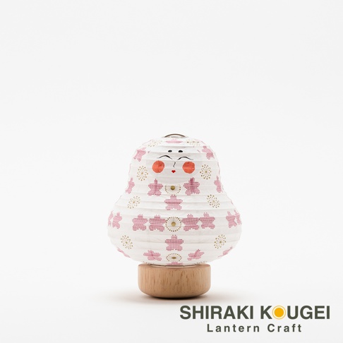 一個白色紙質達摩不倒翁迷你提燈，上面印有粉紅色櫻花圖案和金色圓形裝飾，臉部有紅色圓形臉頰、黑色眼睛和嘴巴，底部為木質底座。