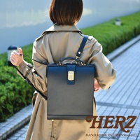 黑色皮革製成的杜勒斯商務後背包，方形設計，配有金色鎖扣和提把，展示於身穿卡其色風衣的模特兒肩上。