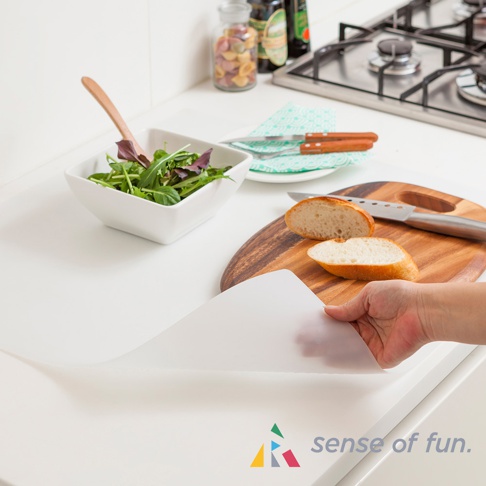 一張白色的耐高溫料理台保護墊，展示在廚房料理台上，旁邊有沙拉碗、麵包、刀具及烤爐。