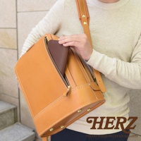 一個使用淺棕色皮革製成的立體商務後背包，背包正面有一個拉鍊口袋，裡面放著一個深棕色皮夾。背包採用手工縫線和黃銅鉚釘裝飾，展現精緻工藝。