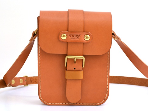 職人手作皮革經典皮扣單肩包，採用棕褐色皮革製成，配有金屬皮扣和品牌標誌。