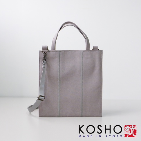 淺灰色棉帆布托特包，兩側有垂直縫線，配有提把和可拆卸肩帶，包包正面左側有銀色金屬扣環。