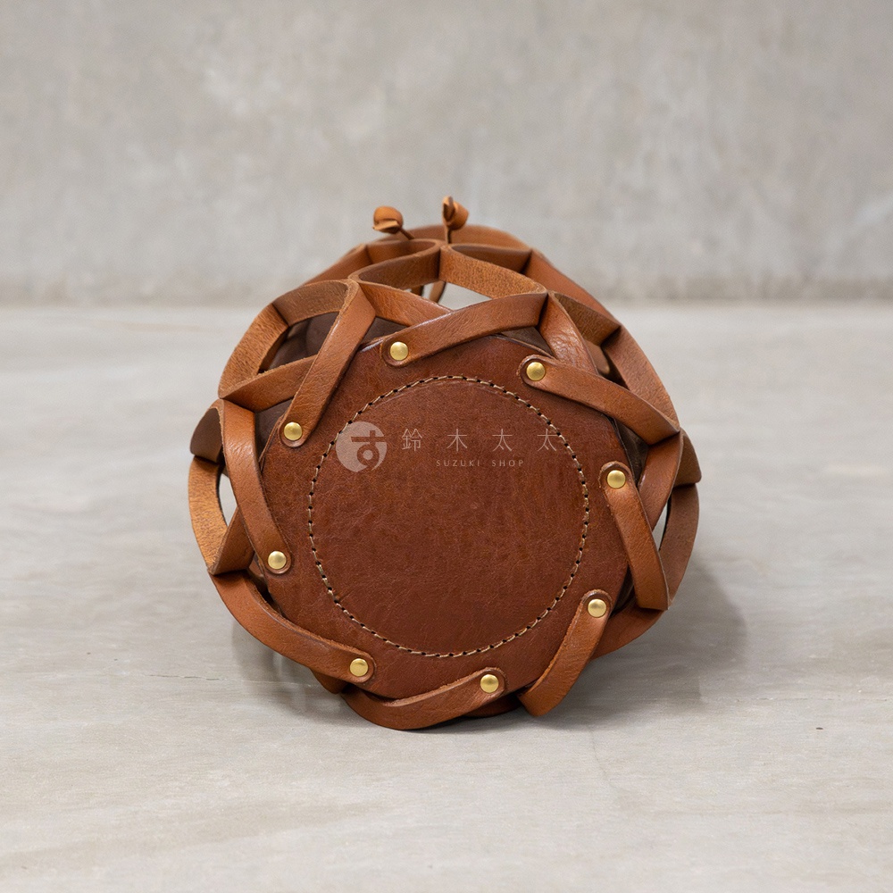 一個以圓形底部和編織皮革側面設計的棕色皮革水桶包，包身由多條皮革條狀物編織而成，側邊有金色鉚釘裝飾，底部中央有SUZUKI SHOP的商標。