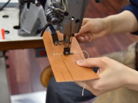 職人以縫紉機正在縫製淺棕色皮革長夾，皮革表面有三條燙金線條，縫線為白色。