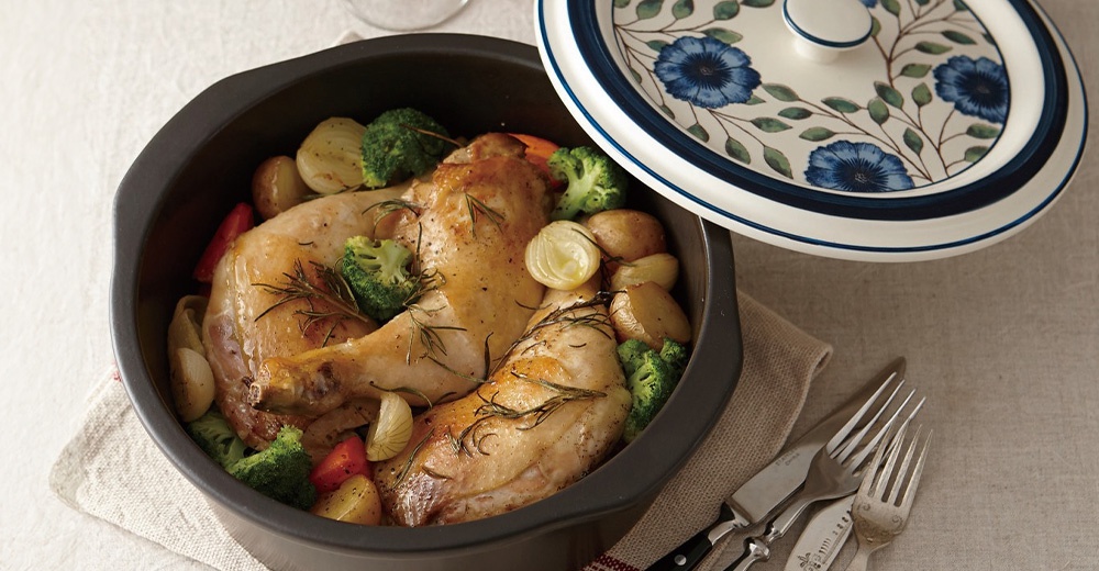 一道裝在黑色圓形烤盤中的烤雞，周圍擺放著烤馬鈴薯、花椰菜和洋蔥，烤盤蓋上有藍色花朵的裝飾。