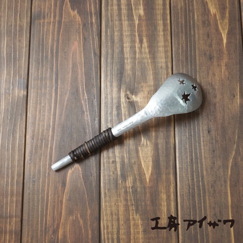 銀色鋁製中型瀝水湯勺，勺頭為錘目紋，帶有星形鏤空，手柄部分纏繞著深棕色繩線，置於木紋背景上。