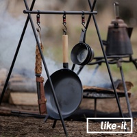 黑色鐵製平底鍋，配有木柄，懸掛在金屬支架上，旁邊有兩個金屬湯勺。背景有營火和傳統水壺。