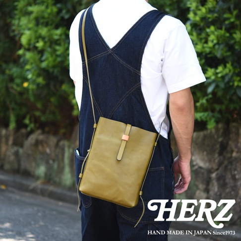 一位背著日本HERZ職人手作橄欖綠皮革直式單肩包的男子，包款呈現極簡俐落的扁平結構，中央設有獨特皮帶釦設計，搭配深藍色丹寧吊帶褲，展現舒適休閒的日系風格，背景為自然綠色植物。