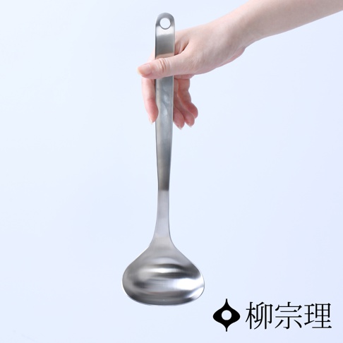 這是一張柳宗理不鏽鋼湯勺M的產品展示圖，畫面中央為一隻手拿著銀色霧面質感的不鏽鋼湯勺，湯勺設計簡約且線條流暢，柄端設有圓孔方便懸吊，右下方印有柳宗理品牌標誌與文字，背景為乾淨的純白色。