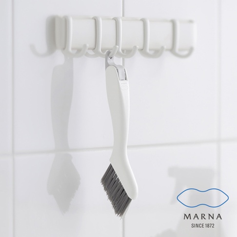 白色塑膠手柄的縫隙清潔刷，刷毛為灰色，刷頭傾斜，掛在白色掛架上，背景為白色磁磚牆面。