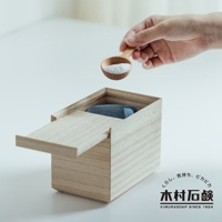 手持木製湯匙，舀取白色洗衣粉，倒入淺色木盒中。木盒蓋子部分開啟，盒內可見藍色包裝的產品。