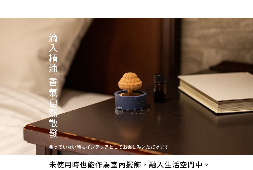 在深色木質床頭櫃上，放置著一個精油擴香座，其底部為藍色圓形底座，上方裝飾著一個木紋清晰的杉木小樹造型。旁邊擺放著一瓶深色精油瓶與一本簡約書籍，整體呈現溫馨舒適的室內氛圍，文案強調精油透過擴香樹自然散發香氣，且未開啟使用時亦可作為精緻的室內擺飾。