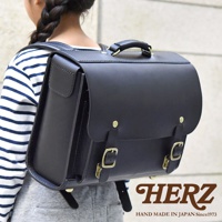 黑色皮革橫式書包，配有金屬扣環和提把，由一位穿著條紋上衣的小女孩背著。