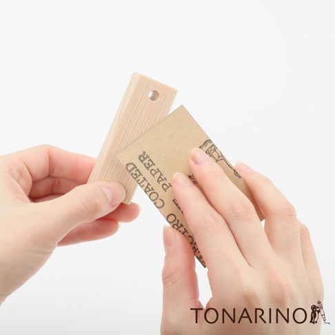 雙手正拿著一塊淺木色、帶有紋理的檜木條，及一張牛皮紙砂紙。檜木條一端有一個小圓孔。
