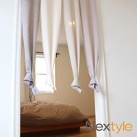 神戶異國風四開式門簾，由淺灰色、米白色布料製成，垂掛於門框，底部綁成結。門簾後方可見臥室內床鋪及窗戶。