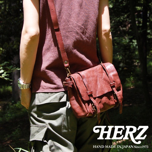 身著酒紅色上衣及卡其色工裝褲的人，斜挎一個紅棕色復古皮革郵差包，包包為水洗加工，配有兩條可調節皮帶扣。