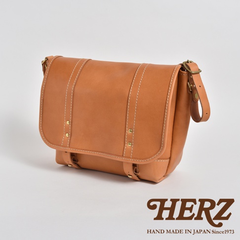 職人手作皮革復古郵差包，以天然棕色皮革製成，搭配白色車線及金色金屬扣飾，設計簡約實用。