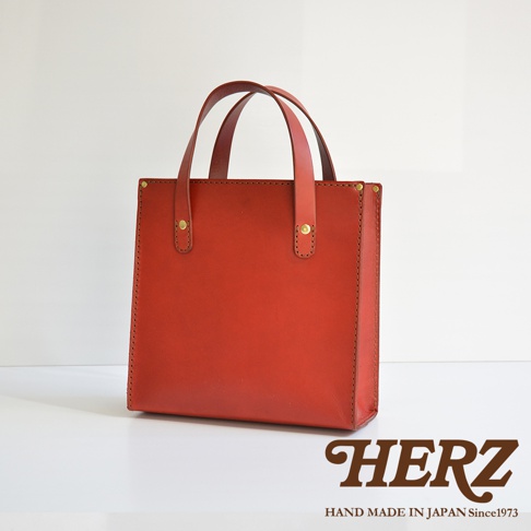 紅色職人手作皮革直式托特包，簡約方正設計，附有兩條皮革提把，包身側邊與頂部邊緣以縫線裝飾，並以金色鉚釘固定提把。