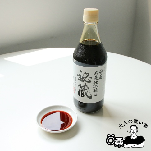 一個裝有深褐色醬油的玻璃瓶，瓶身貼有白色標籤，印有黑色日文漢字「秘藏」及其他日文說明。瓶蓋為淺黃色。旁邊有一個白色小碟，碟中盛有少量醬油。