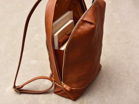 一個簡約褶皺風的棕色皮革後背包，部分拉鍊敞開，露出內部裝有幾本書籍。背包有一條皮革肩帶。