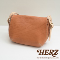 棕色皮革製成的郵差包，帶有可調節的淺色皮革肩帶和金屬扣環。包身有自然的皮革紋理和手工縫線細節。