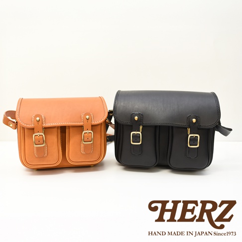左側是棕色皮革郵差包，右側是黑色皮革郵差包，兩款包均有雙皮帶扣和雙前袋，底部印有HERZ HAND MADE IN JAPAN Since1973字樣