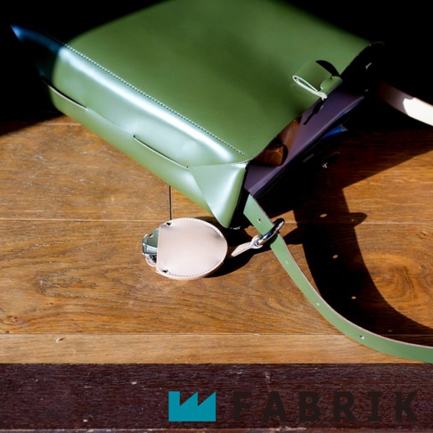 FABRIK 品牌綠色皮革方形包，側面掛有一個淺棕色皮革圓形鑰匙圈，放置在木質桌面。