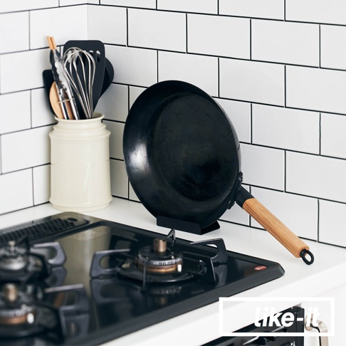 黑色直火專用萬能調理鐵鍋，鍋身為鐵質，附有木質手柄，置於黑色鍋架上，旁邊有白色陶瓷鍋具收納筒，筒內裝有打蛋器、鍋鏟等廚房用具。
