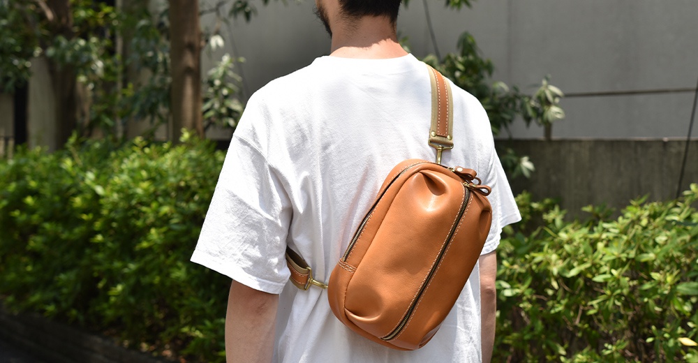 一個身穿白T恤的男子，背著一個棕褐色的皮質斜背包，包包位於他身後，並橫跨在他的肩膀上。